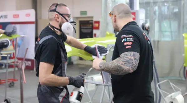 Dwóch mężczyzn w maskach i rękawiczkach pracuje nad zielonym elementem, sprawdzając, czy na podkład epoksydowy można kłaść lakier.