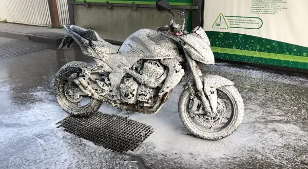 Motocykl pokryty pianą na myjni. Widać instrukcję, jak myć motocykl, z zaleceniem zachowania bezpiecznej odległości.
