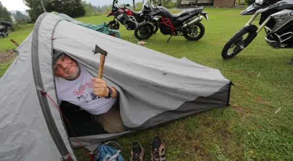Mężczyzna w namiocie z siekierką, obok motocykle. Warto zabrać na wyprawę motocyklową namiot, buty i siekierkę.