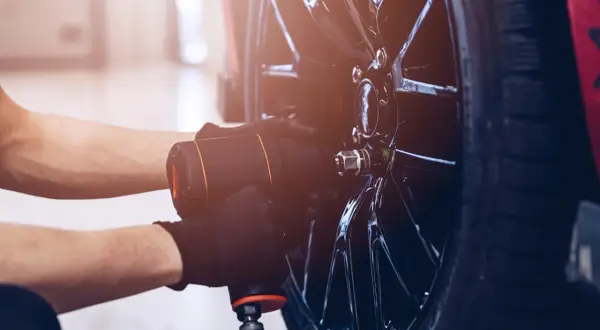 Mechanik w czarnych rękawiczkach używa klucza pneumatycznego do odkręcania śrub w feldze, przygotowując się do wymiany jednej opony.