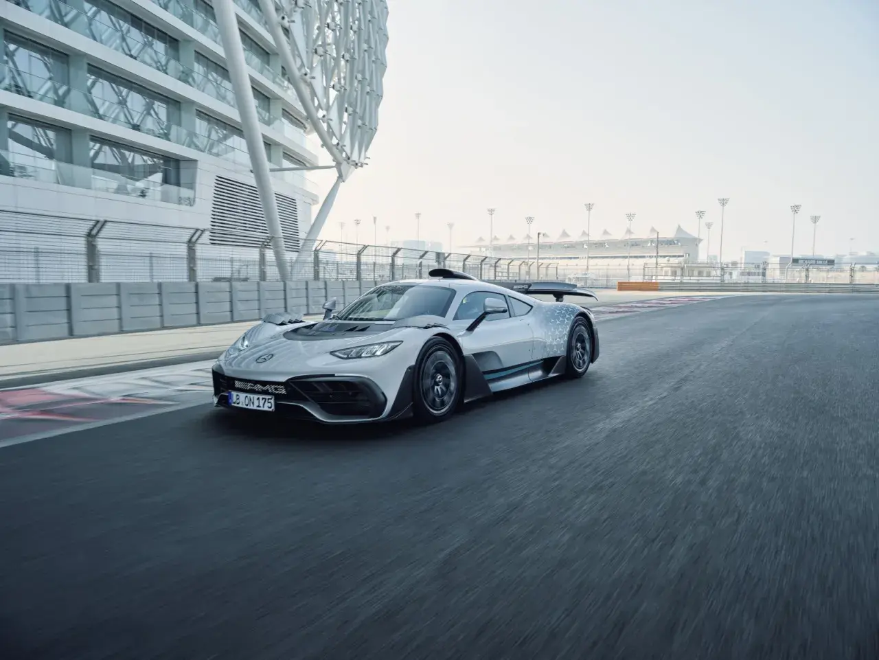 Srebrny Mercedes-AMG Project One pędzi po torze wyścigowym, obok nowoczesnej architektury.