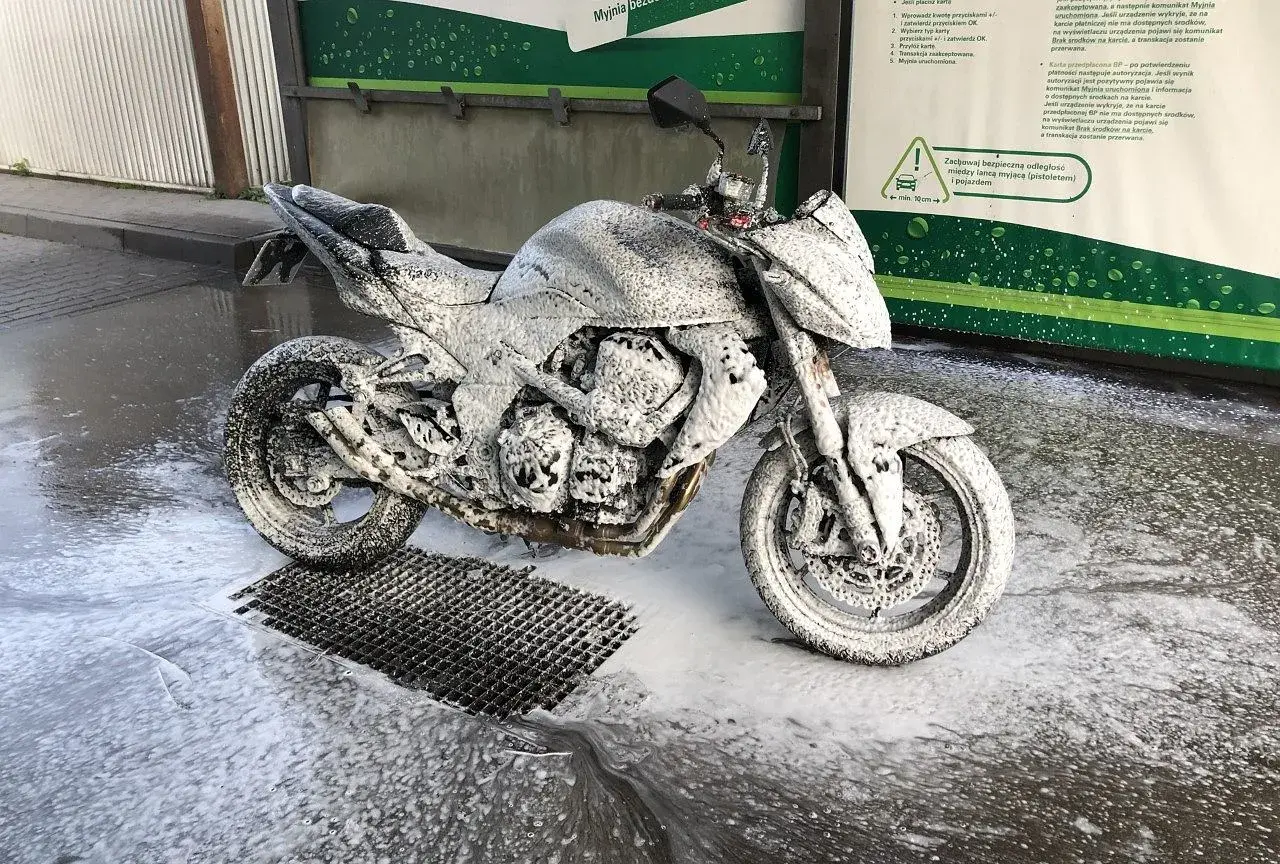 Motocykl pokryty pianą na myjni. Widać instrukcję, jak myć motocykl, z zaleceniem zachowania bezpiecznej odległości.