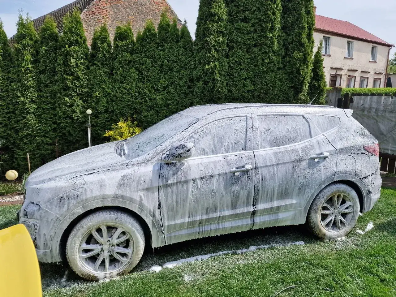 Samochód pokryty obfitą pianą aktywną, gotowy do mycia na tle zielonego ogrodu i domu.