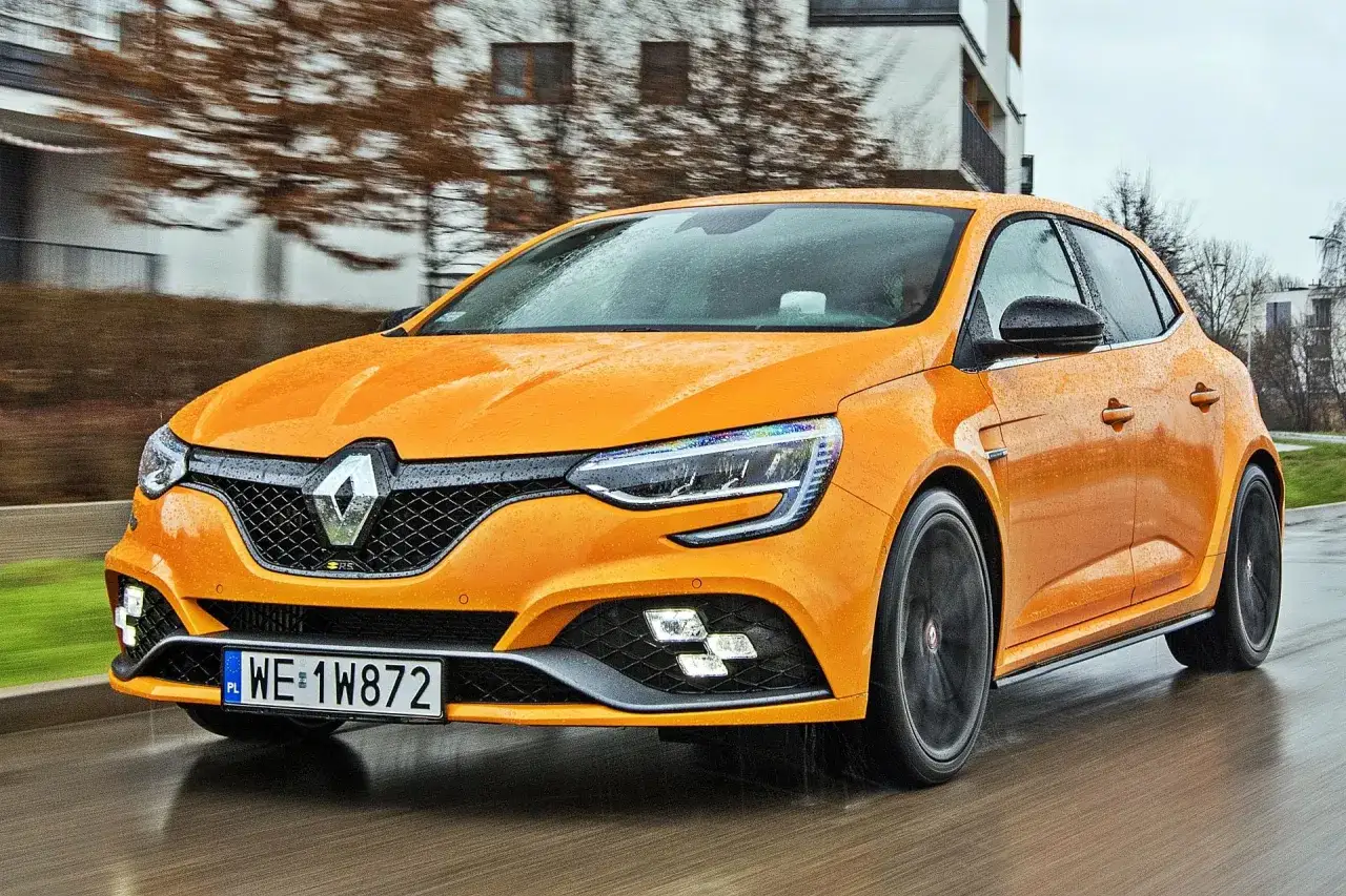 Pomarańczowy Renault Megane RS w deszczu, z kroplami wody na karoserii, pędzi po mokrej drodze.