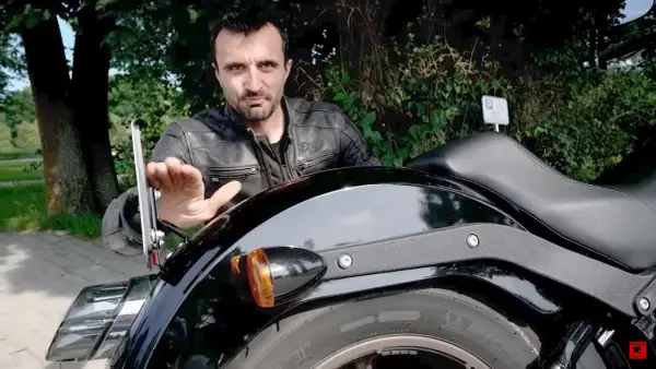 Mężczyzna w skórzanej kurtce pokazuje mocowanie tablicy motocyklowej.